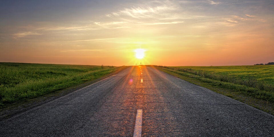 a road with grass on the side and the sun setting